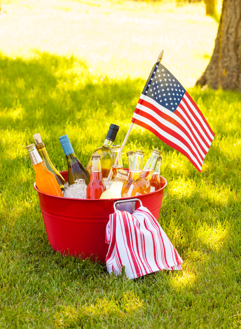 Cold drinks wine, beer and soda on ice in red bucket pail tub with American flag  on green grass. Summer outdoor backyard Fourth of July 4th garden party picnic.