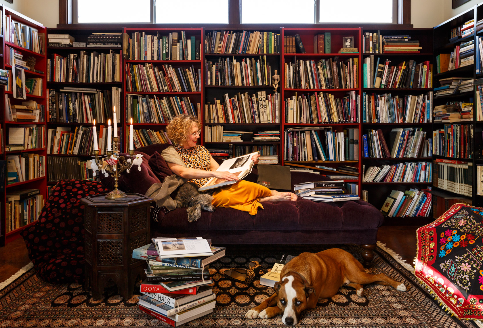 Happy, smiling, creative middle aged woman with her pet dogs relaxing reading a book in her unique home library interior